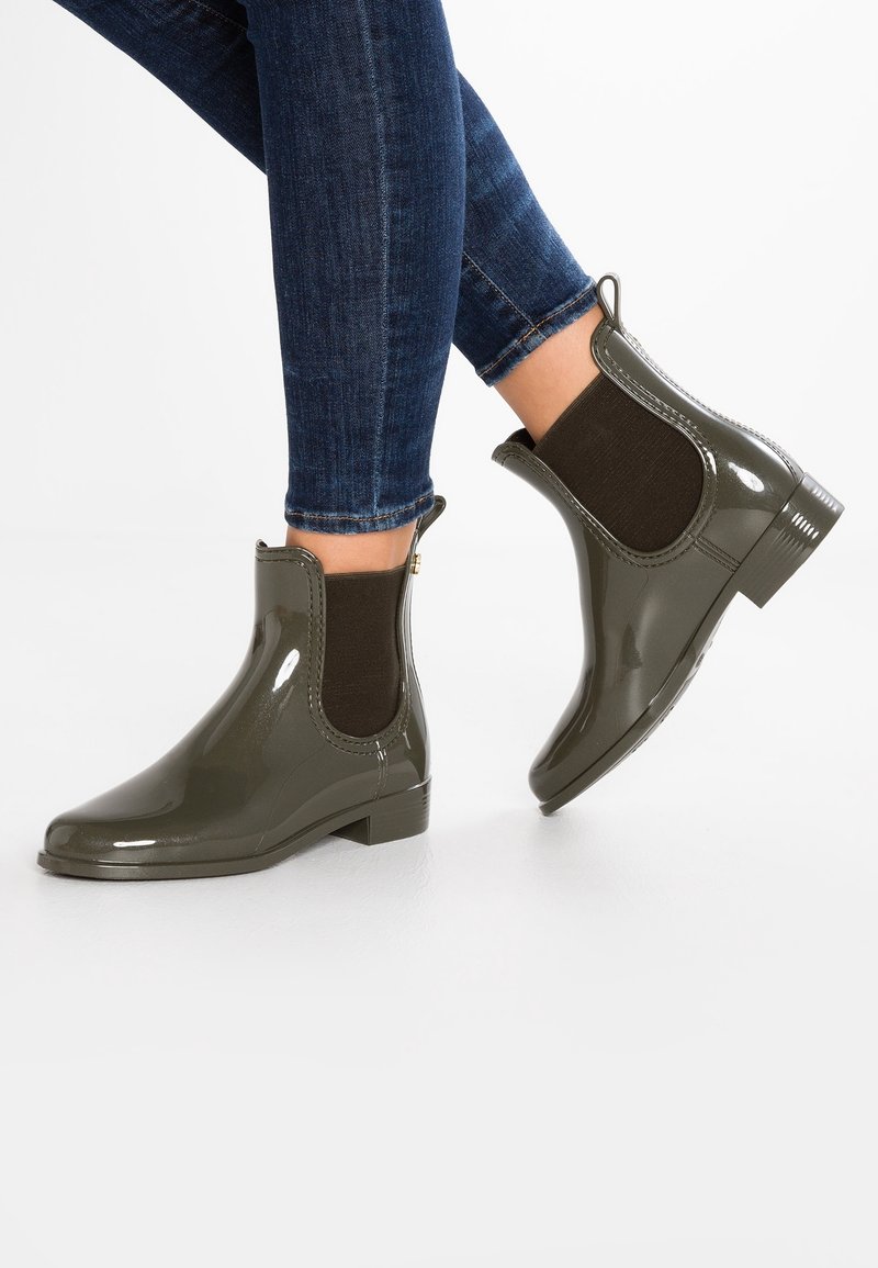 Persona vistiendo botas de lluvia brillantes de color verde oscuro hasta el tobillo y jeans ajustados azules, contra un fondo blanco liso.
