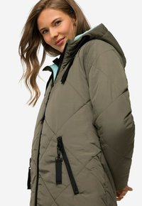 Giubbotto imbottito trapuntato verde oliva con cappuccio azzurro chiaro, cerniere nere e lacci. Tessuto strutturato e motivo a rombi prominente.