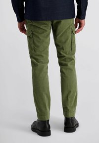 Man in olijfgroene corduroy cargobroek en zwarte leren laarzen, staand tegen een effen lichte achtergrond.