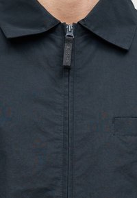 Σκούρο μπλε ζακέτα με φερμουάρ, με μεγάλο γιακά, μπροστινό φερμουάρ και μια μικρή τσέπη στην αριστερή πλευρά. Το υλικό φαίνεται ελαφρύ και υφασμάτινο.