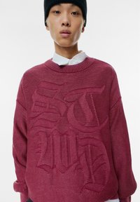Maglione oversized a maglia in bordeaux con design testuale in rilievo. Presenta un collo rotondo a costine e spalle ribassate. Dettaglio in tessuto testurizzato.