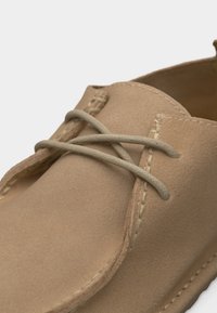 Chaussure décontractée en daim marron clair avec bout arrondi, coutures visibles et lacets beige noués en un nœud lâche.