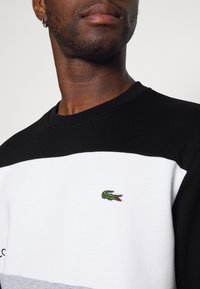 Uomo che indossa una felpa a blocchi di colore nera, bianca e grigia con un piccolo logo di coccodrillo verde sul petto e un orecchino ad anello in argento.