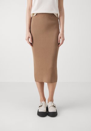 Vrouw draagt een beige geribde midi-rok en witte loafers met zwarte zolen, staand tegen een eenvoudige lichte achtergrond.