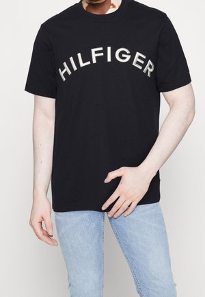 Man wearing black short-sleeve T-shirt with white "HILFIGER" text across the chest and light blue jeans, standing against plain background.