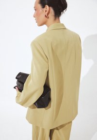Prešívaný béžový blejzer s pevnými ramennými vycpávkami, doplnený čiernou koženou kabelkou. Hladká textúra a minimalistický dizajn.