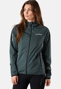 Veste légère de couleur vert foncé, avec une fermeture éclair complète, une capuche et des poches latérales. Possède des accents réfléchissants et une texture lisse et résistante à l'eau.