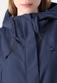 Giacca impermeabile blu navy con colletto alto, doppio bottone e cordini elasticizzati. Presenta una texture liscia e cuciture minime.