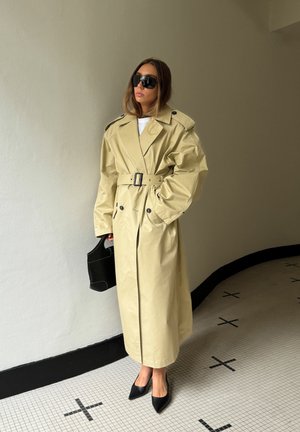Beige trenchercoat, jossa on vyötä ympäröivä vyö, suuret kaulukset ja kaksirivinen malli. Musta käsilaukku ja kärjelliset mustat korkokengät ovat myös näkyvissä.