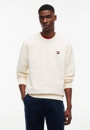 Mężczyzna w kremowej bluzie Tommy Jeans i granatowych spodniach, stojący z jedną ręką w kieszeni na jednolitym białym tle.