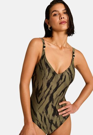 Femme portant un maillot de bain une pièce à motifs vert olive et noir avec des bretelles fines et des accents de perles dorées, posant avec la main sur la hanche.