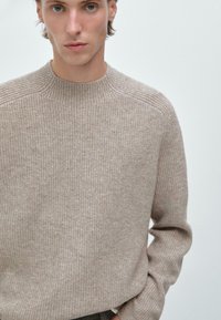 Beige ribbet striktrøje med rund hals, med lange ærmer og en tekstureret overflade, vist foran en ensfarvet baggrund.