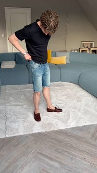 Hnedé kožené mokasíny s šnúrkami, denimové šortky a čierna košeľa s krátkym rukávom, stojace na texturovanom svetlom koberci.