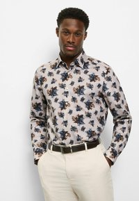 Herren-Hemd mit Knopfleiste in hellem Beige mit blauem und braunem Blumenmuster. Verfügt über einen Kragen und lange Ärmel, kombiniert mit einem dunklen Gürtel und cremefarbenen Hosen.