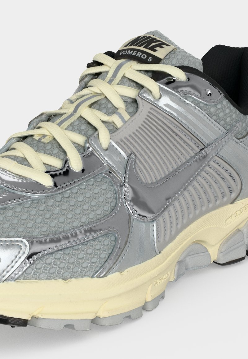 Zapatilla Nike Vomero 5 en gris y plata con cordones color crema y suela acolchada, con paneles superpuestos texturizados y brillantes.