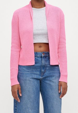 Le modèle porte un cardigan en maille rose par-dessus un haut court gris et un jean bleu.