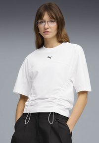 Νεαρή γυναίκα με γυαλιά, ντυμένη με λευκό πουκάμισο Puma με κορδόνια και μαύρο παντελόνι, με τα χέρια στις τσέπες, μπροστά σε γκρι βαθμιδωτό φόντο.