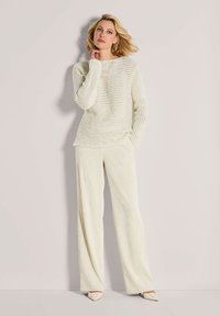 Frau mit blonden Haaren, die einen cremefarbenen Strickpullover, weite beigefarbene Hosen und spitze beiger Schuhe trägt und vor einer schlichten hellen Wand steht.