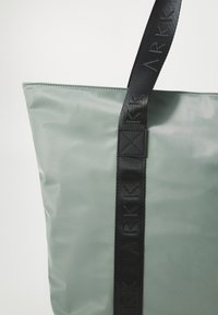 Zaļa neilona somiņa ar melnu austu siksnu, kurai ir izspiestā logo. Gluda struktūra, taisnstūrveida forma un augšējais aizvērums.