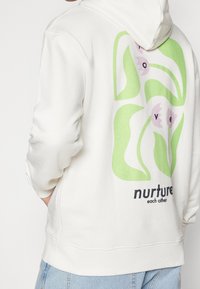 Sweat à capuche crème avec un grand motif de feuilles vertes et de fleurs roses au dos. Texte "prenez soin les uns des autres" sous le dessin.