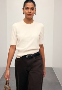 Pull crème à manches courtes avec un col côtelé, associé à un pantalon marron taille haute, accentué par une ceinture noire et portant un sac.