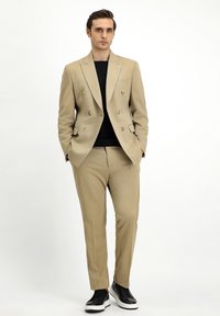 Blazer beige doppiopetto con bottoni dorati, abbinato a pantaloni beige su misura e sneakers nere. Tessuto liscio, vestibilità moderna.