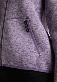 Fialová bunda na zip s texturovanou tkaninou s flíčky, vybavená boční kapsou a značkovou cedulkou, doplněná hladkým lemováním podél švů.