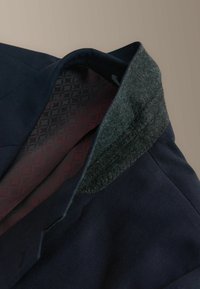 Marineblazer mit grünem Filzkragen, verziert mit einem gemusterten burgunderroten Futter. Verfügt über strukturierte Schultern und einen texturierten Stoff.