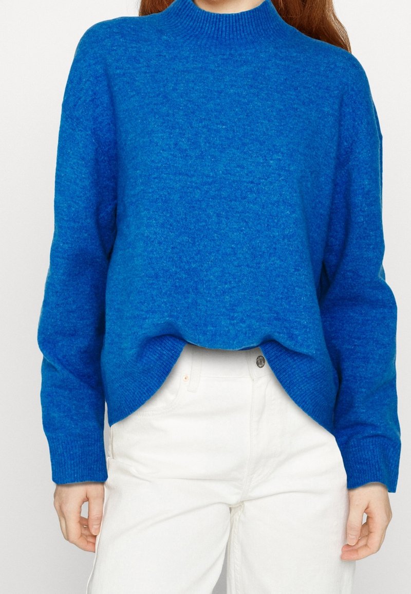 Personne portant un pull en tricot bleu vif à manches longues associé à un pantalon blanc taille haute sur un fond uni.