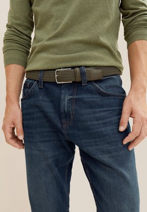 Ceinture tissée élastique vert olive, dotée d'une boucle en métal argenté, assortie à un jean en denim bleu foncé avec un léger délavage et des accents de couture rouge.