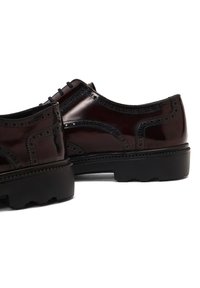 Zapatos de cuero marrón oscuro con detalles de brogue, puntera redondeada, suela negra contrastante y cordones planos, que muestran un acabado pulido.