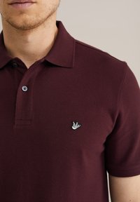 Burgundin punainen polo-paitaa, jossa on teksturoitu kangas, kolmonen nappilista ja brodeerattu lintulogolla vasemmassa rinnassa.