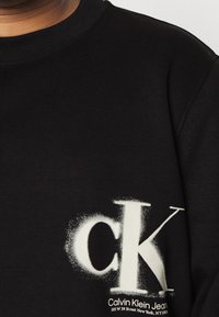 Svart sweatshirt med en stor vit "ck"-logotyp, med sprayad effekt och mindre varumärkestext i nedre vänstra hörnet.