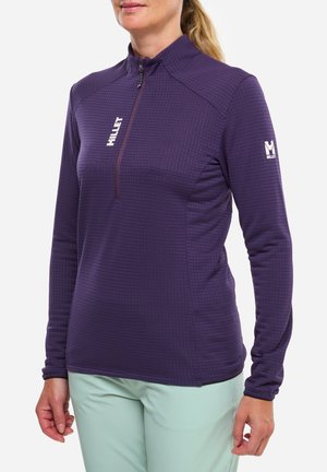 Femme portant un haut violet texturé à manches longues avec une demi-fermeture éclair et un logo blanc sur la poitrine et la manche, associé à un pantalon vert clair.