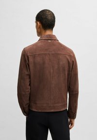 Giacca in suede marrone con colletto, cuciture visibili e polsini abbottonati. La vista posteriore mostra un design pulito e un discreto logo sul collo.