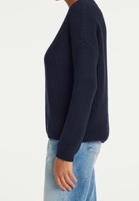 Personne portant un pull en maille texturée bleu foncé et un jean bleu clair, vue de profil sur un fond uni.