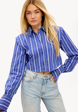 Chemise bleue coupe courte avec des rayures verticales blanches, fermeture à boutons, manches longues avec larges poignets, associée à un jean en denim bleu clair.