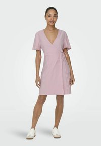 Robe portefeuille rayée rose et blanche avec des manches courtes, décolleté en V et un détail à nouer à la taille. Associée à des sneakers blanches.