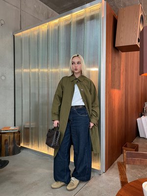 Személy szőke hajjal, olívazöld kabátot, fehér inget, széles sötét farmert és bézs cipőt viselve, sötét szövött kézitáskát tartva beltéren.