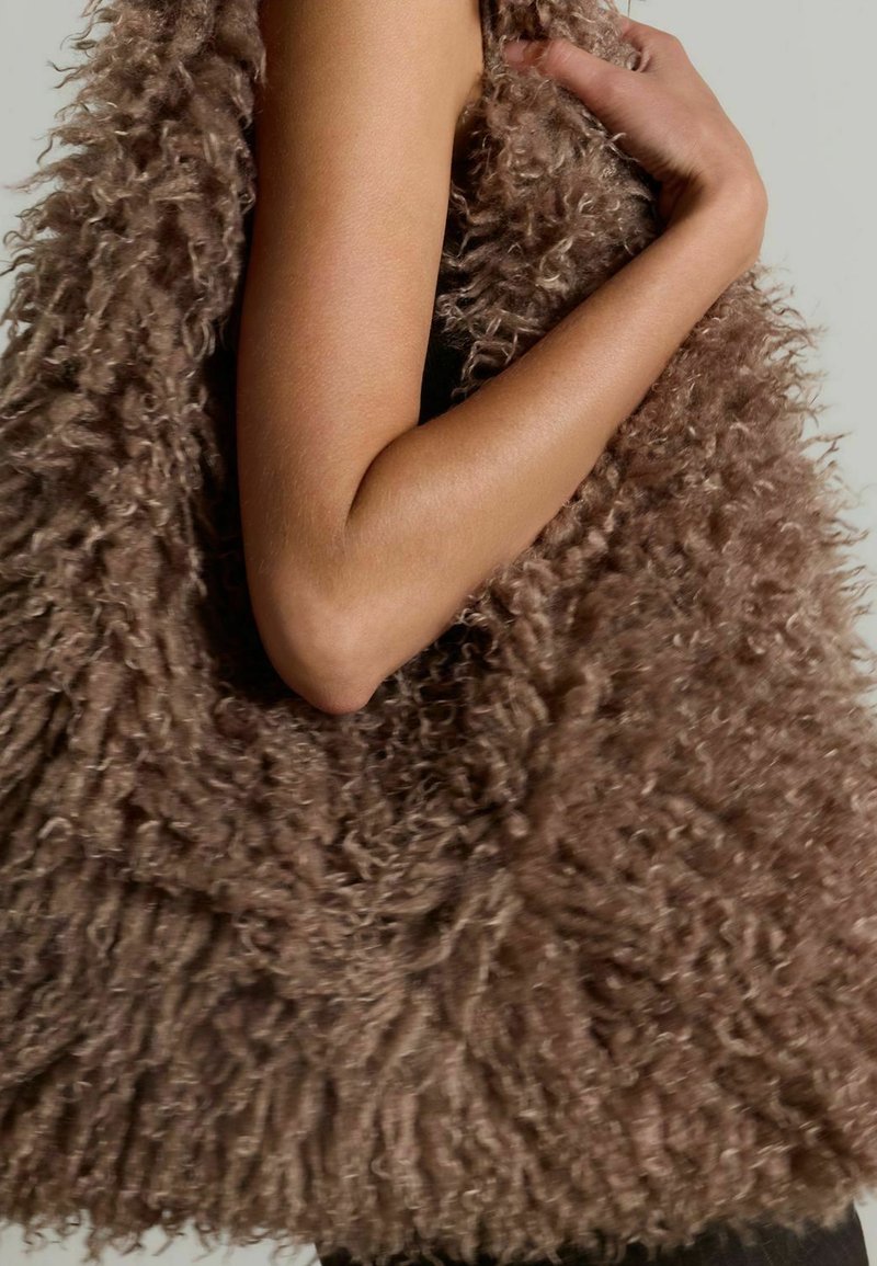 Sac fourre-tout marron duveteux avec une surface texturée et hirsute, tenu par le bras d'une personne. Le design présente des bords doux et effilochés ainsi qu'une forme arrondie.