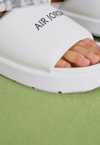 White slide sandals with a wide strap featuring "AIR JORDAN" logo. Smooth texture, rubber sole, and minimalist design. Close-up view.