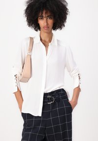 Camisa blanca de botones con puños abotonados que presenta recortes decorativos. Combinada con pantalones de cuadros azul marino y un bolso beige.