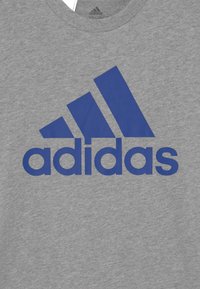 Camiseta de algodón gris con un gran logo azul de Adidas. El diseño incluye tres franjas diagonales sobre el nombre de la marca.