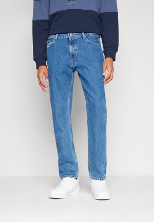 Person trägt gerade geschnittene blaue Jeans, weiße Sneaker und ein marineblaues Sweatshirt mit Farbblockmuster und steht vor einem schlichten Hintergrund.