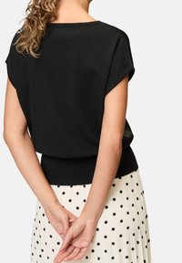 Czarny dzianinowy top z krótkimi rękawami i prążkowanym paskiem w talii, noszony na białej spódnicy w czarne grochy. Widok od tyłu.