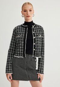Musta-valkoinen tweed-takki, jossa on fringes-reunus, pyöreä kaula-aukko ja nappikiinnitys. Yhdistetty samansävyiseen ruutuhameeseen.