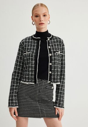 Musta-valkoinen tweed-takki, jossa on fringes-reunus, pyöreä kaula-aukko ja nappikiinnitys. Yhdistetty samansävyiseen ruutuhameeseen.