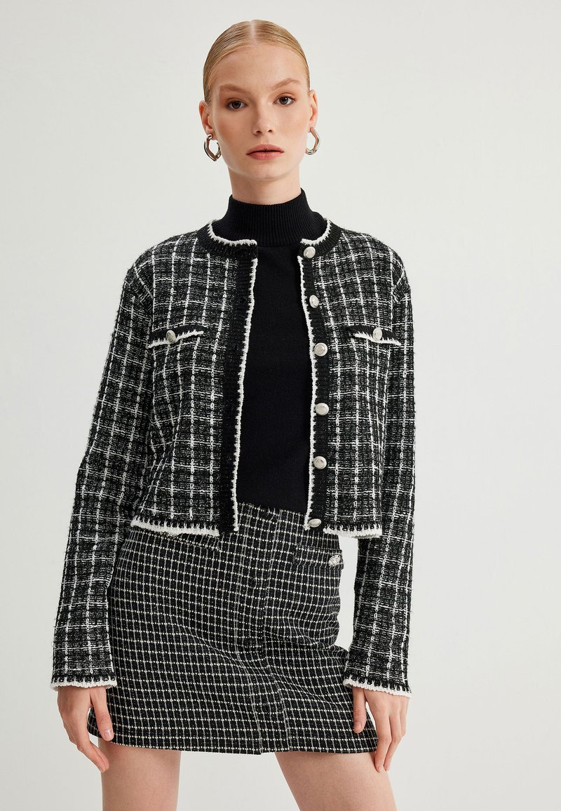 Schwarz-weißer Tweed-Jacke mit Fransenbesatz, rundem Ausschnitt und Knopfverschluss. Kombiniert mit einem passenden karierten Rock.