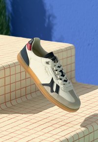 Sneaker blanche avec des accents noirs, gris et rouges flottant au-dessus de marches en carrelage beige contre un fond mural bleu vif.