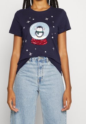 Kobieta w granatowym t-shircie z brokatowym pingwinem w projekcie śnieżnej kuli, do tego jasnoniebieskie, wysokie jeansy.